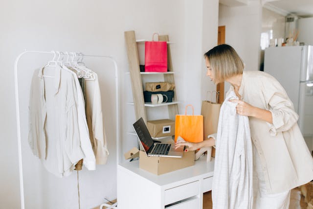 A person in a light-colored outfit holds a jacket and an orange shopping bag while standing near a white desk with an open laptop and a cardboard box, suggesting online shopping. A clothing rack with white shirts, a shelf with pink and brown bags, and a refrigerator are visible in the background, indicating a home environment.