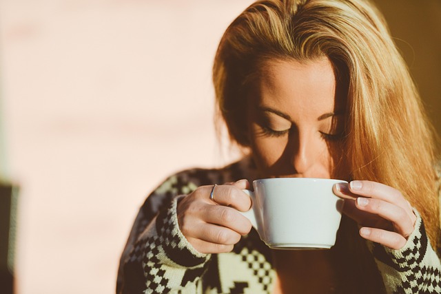 A woman with long blonde hair holding a white cup close to her face, appearing to enjoy the warmth and aroma, with a soft-focus background and warm lighting.